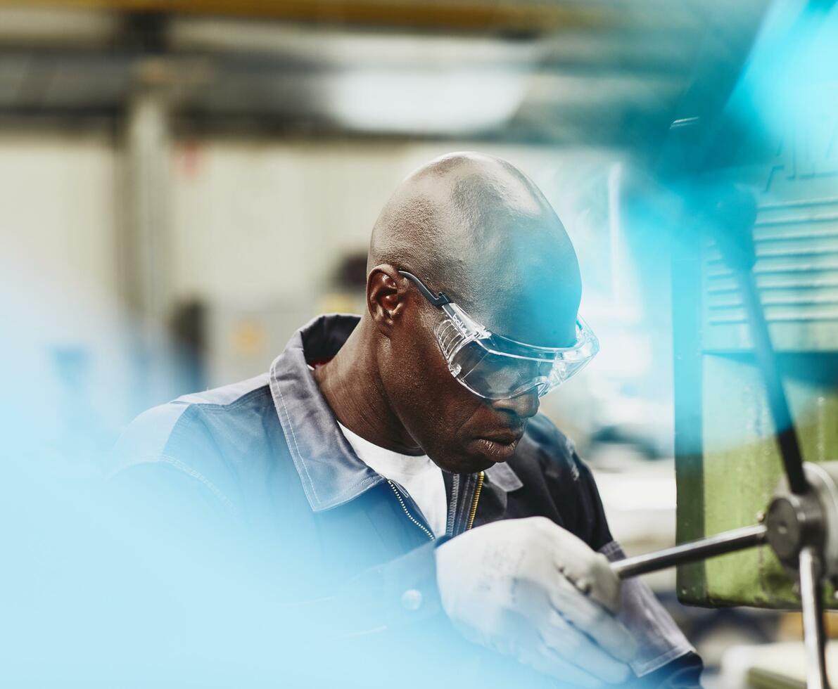 Factory worker operating a column drill. African-American man wearing safety goggles. Primary color blue.