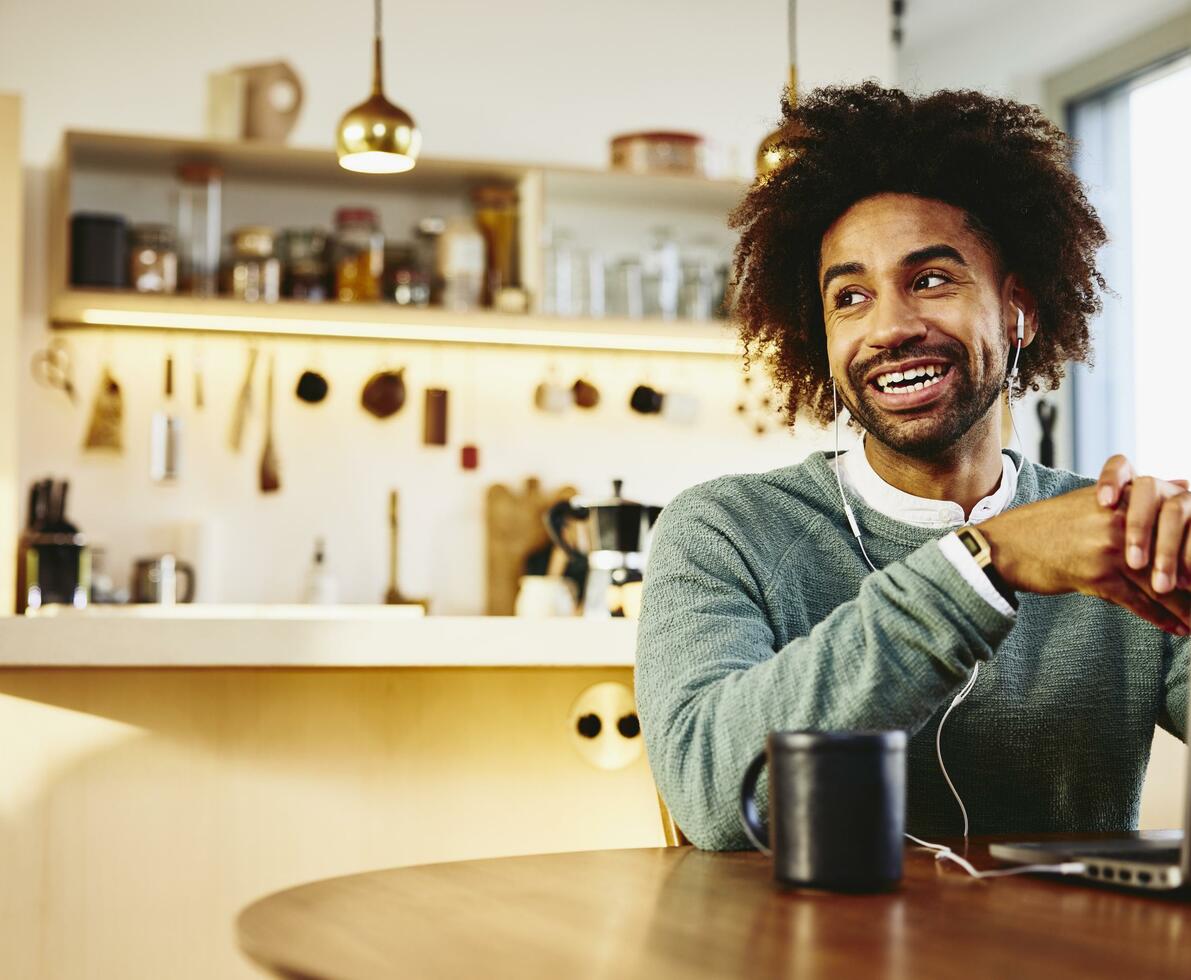 Smiling man sitting at his dining table with a drink and his laptop. Looking away. Kitchen in the background. Smiling man sitting at his dining table with a drink and his laptop. Looking away. Kitchen in the background.