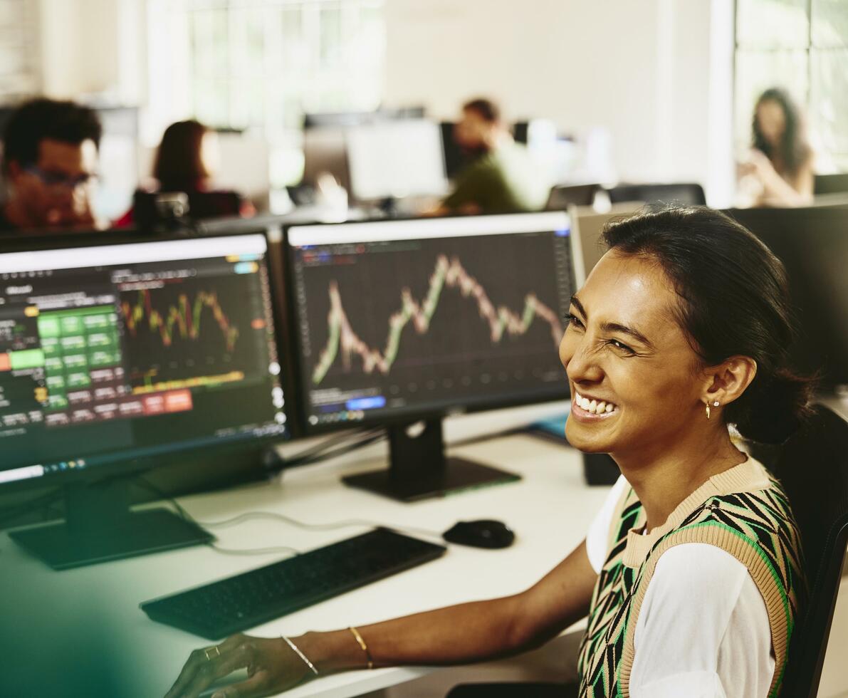 Woman smiling while sitting behind her desk, screens displaying financial information.