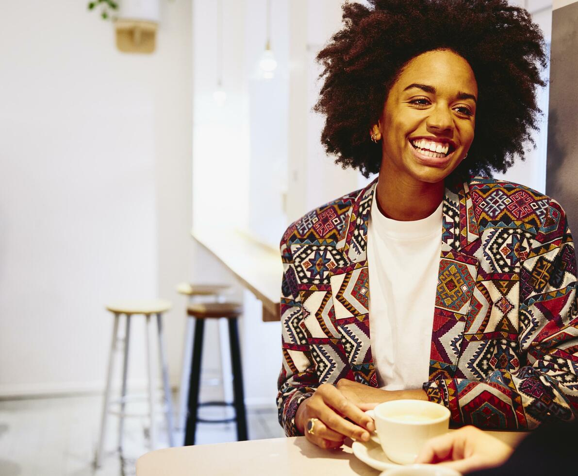 Smiling woman sitting at a table with coffee, smiling at someone.