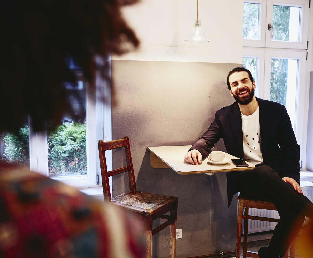 Man sitting at table with a coffee, smiling to someone